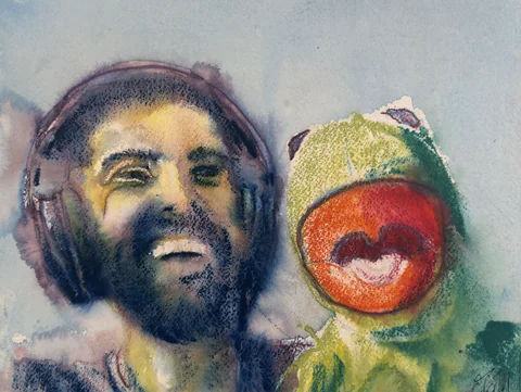 A laughing man wearing headphones beside a green frog puppet