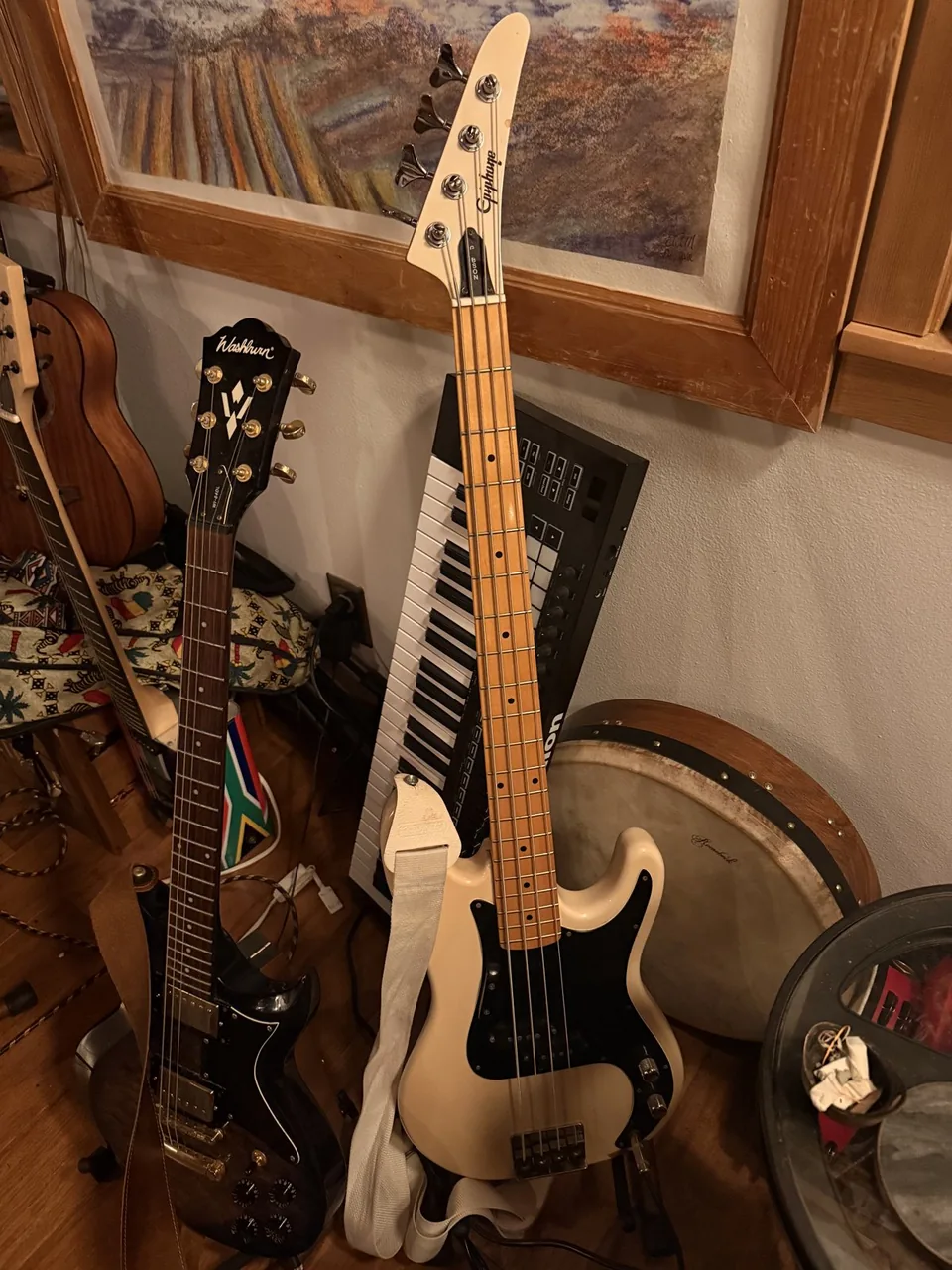 A cream Epiphone bass guitar with a black pickguard and maple neck, leaning against the wall next to a Washburn electric guitar