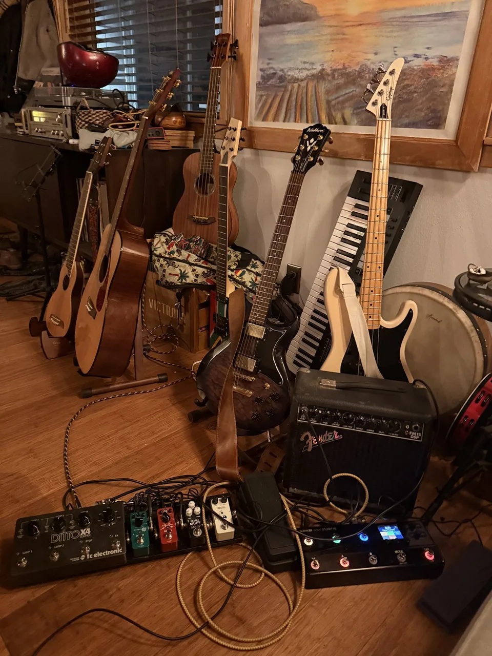 My music corner: acoustic guitars, a Washburn electric, a cream Epiphone bass, a keyboard and a hand drum leaning against the wall, with a Fender amp and a pedalboard on the floor